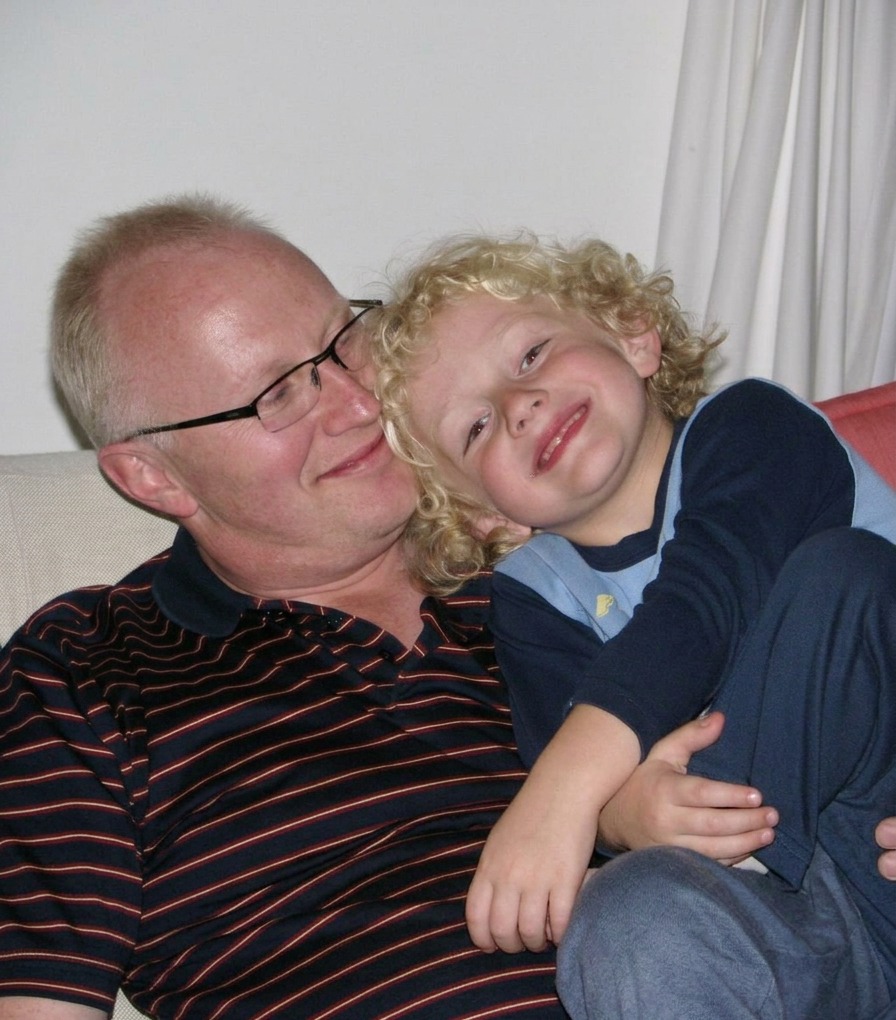 Father resting cheek-to-cheek with his curly-haired young son on the couch