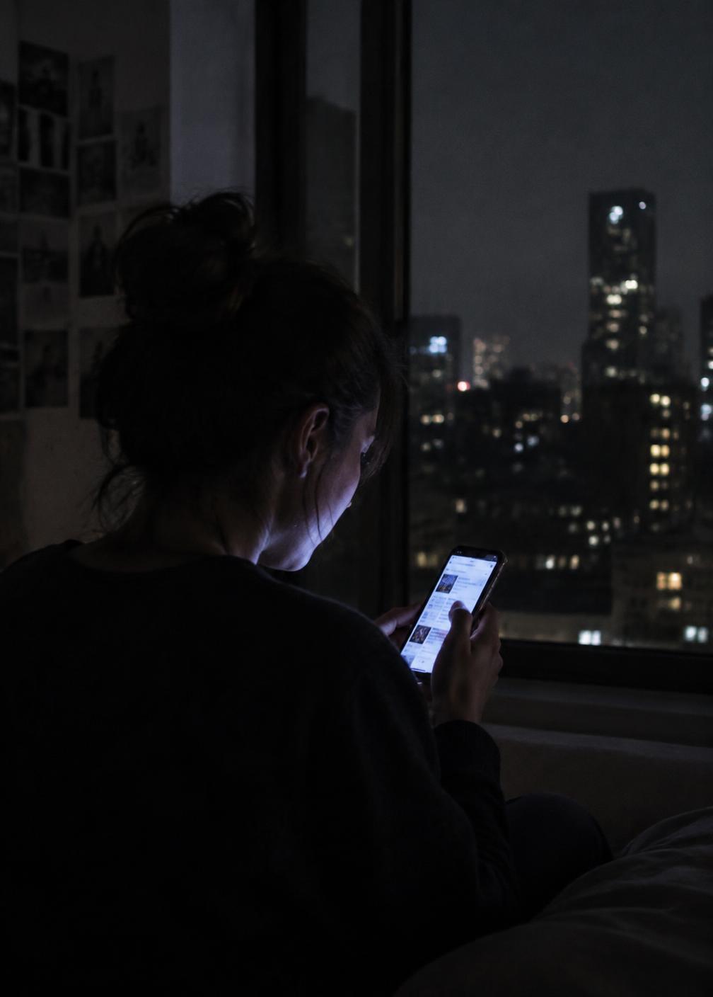 A young woman scrolling a phone in a dark room, her face lit by the screen